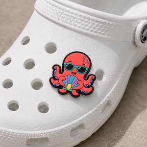 May include: A white foam clog shoe featuring a decorative rubber charm shaped like a red octopus wearing teal sunglasses and holding a purple and blue seashell. The octopus has purple suction cups on its tentacles and a black outline.