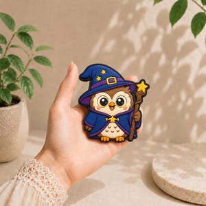 May include: A rubberized patch featuring a cartoon owl dressed as a wizard in a dark blue robe and matching pointed hat with yellow stars. The owl holds a brown wooden staff topped with a yellow star and has large eyes and a light brown belly.