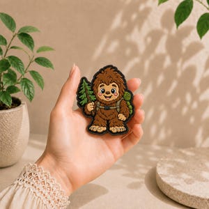 Könnte beinhalten: Ein brauner Bigfoot-Aufnäher im Cartoon-Stil, der eine lächelnde Kreatur mit rosa Wangen zeigt, die einen grünen Rucksack trägt und einen kleinen grünen Tannenbaum hält. Der Aufnäher hat einen dicken schwarzen Rand und ein detailliertes Design.