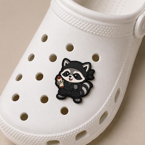 May include: A white foam clog shoe featuring a decorative rubber charm shaped like a cartoon raccoon dressed in a black ninja outfit. The raccoon character is holding a small tan scroll and is attached to one of the circular ventilation holes.