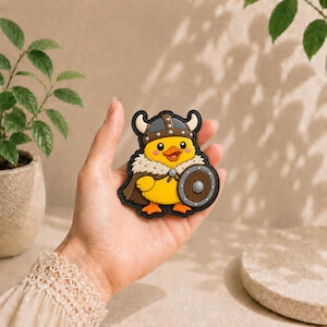 Peut inclure: Un écusson en forme de canard jaune portant un casque gris à cornes, une cape marron avec une bordure en fourrure blanche et un bouclier rond marron. Le canard a des pattes et un bec orange, avec un contour noir autour du motif.