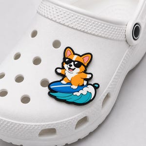 Op de afbeelding: Een witte rubberen klomp met een decoratieve bedel van een cartoon corgi met een zwarte zonnebril die op een blauw surfboard over een turquoise oceaangolf surft, bevestigd aan de bovenkant van het witte schoeisel.