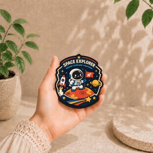 May include: A circular patch featuring a cartoon astronaut standing on a red planet, holding a red flag with a rocket icon. The design includes a rocket, a yellow planet, stars, and the text SPACE EXPLORER DREAM EXPLORE DISCOVER in white.
