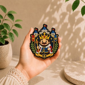 May include: A circular embroidered patch featuring a cartoon king with a crown, beard, and scepter, centered in front of a castle. The design includes red and blue banners, green laurel branches, and gold accents on a dark blue background.