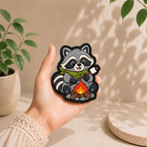 May include: A rubber patch featuring a cartoon raccoon with grey fur, a black eye mask, and a green neckerchief. The raccoon is sitting by a small campfire made of brown logs and orange flames, holding a stick with a white marshmallow on the end.