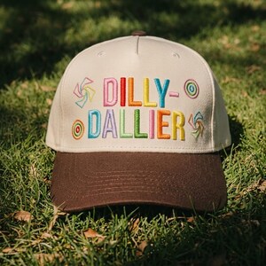 May include: A cream-colored baseball cap with a dark brown brim. The front features embroidered text that reads DILLY-DALLIER in multi-colored letters, accompanied by small, colorful embroidered spiral and circular designs on both sides.