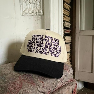May include: A cream-colored baseball cap with a black brim. The front panel features dark purple embroidered text that reads: PEOPLE WHO GET DIARRHEA FROM TACO BELL ARE WEAK AND THEIR BLOODLINE IS WEAK AND HISTORY WILL FORGET THEM.