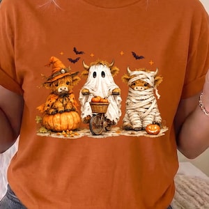 May include: An orange t-shirt featuring a graphic of three highland cows dressed for Halloween. One cow wears a witch hat on a pumpkin, one rides a bicycle as a ghost, and one is wrapped in mummy bandages with a small pumpkin nearby.
