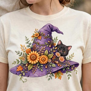 Op de afbeelding: Een crèmekleurig t-shirt met een afbeelding van een paarse heksenhoed versierd met zonnebloemen, herfstbladeren en eikels. Een klein zwart katje met grote ogen kijkt vanachter de rand van de hoed, omringd door groen gebladerte.