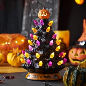 May include: A black ceramic tabletop tree with a glossy finish, decorated with small orange and purple light bulbs. The tree is topped with an illuminated orange jack-o'-lantern and features an orange base with the text Trick or Treat printed on it.