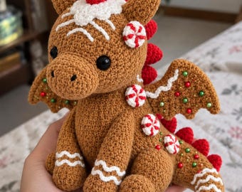 May include: A brown crocheted dragon plush toy decorated like a gingerbread cookie. It features white icing-style embroidery, red and white peppermint candy accents, and small green and red beads on its wings and tail, with black eyes.