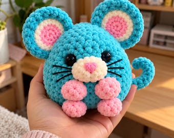 May include: A round, crocheted plush toy shaped like a mouse, featuring bright blue yarn, large ears with white and pink centres, a cream-coloured snout, a pink heart-shaped nose, black plastic eyes, black whiskers, and four small pink feet.