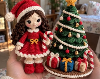 May include: A crocheted doll with brown hair wearing a red dress and Santa hat, holding a candy cane, next to a crocheted Christmas tree decorated with gold bells, red and white spheres, and a yellow star, with three small gift boxes at the base.