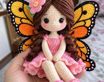 May include: A handmade crochet doll featuring brown yarn hair with antennae, large black eyes, and pink cheeks. The doll wears a pink dress and shoes with flower accents, and has large, detailed orange and black butterfly wings attached.