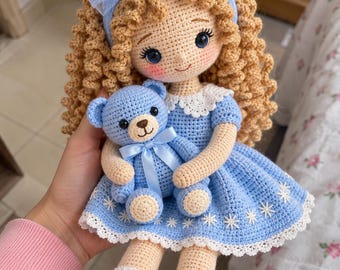 May include: A handmade crochet doll with curly tan yarn hair, wearing a light blue dress with a white lace collar and hem, and a matching blue tulle bow headband. The doll holds a small blue crochet teddy bear with a satin ribbon bow.