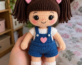 May include: A crochet doll with dark brown hair in pigtails, pink bows, and rosy cheeks. The doll wears a white t-shirt, blue denim-style overalls with a pink heart patch, and pink and white trainers. The doll has large black eyes.