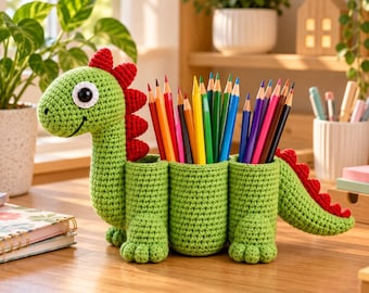 May include: A green crocheted dinosaur pencil holder featuring a long neck, a head with a large white and black eye, and a row of red spikes along its back and tail. The body consists of three cylindrical compartments filled with coloured pencils.