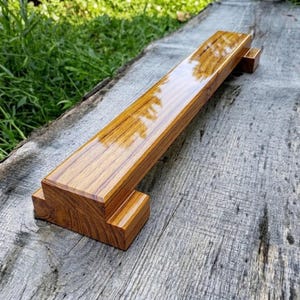 May include: A rectangular wooden handle with a polished, high-gloss finish showcasing a prominent golden-brown wood grain pattern. The handle features a stepped, tiered design at both ends, creating a distinct profile with sharp, clean edges.