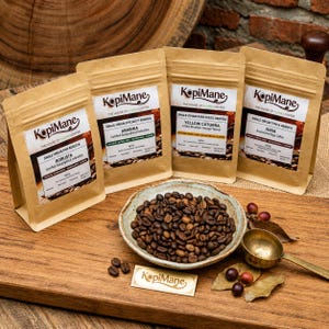 May include: Four brown paper pouches of KopiMane coffee beans labelled Robusta, Arabika, Yellow Caturra, and Juria. A ceramic bowl filled with roasted coffee beans sits in front, accompanied by a metal scoop and a small rectangular brand card.