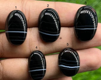May include: Five polished oval black onyx gemstones with distinct white horizontal bands, each labeled with a small black number from one to five. The stones have a smooth, reflective surface and are arranged in a group to display their patterns.