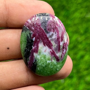May include: An oval-shaped cabochon gemstone featuring a polished surface with a mix of vibrant magenta-red crystal inclusions set against a mottled green and black background. The stone displays a smooth, rounded top and a distinct color pattern.