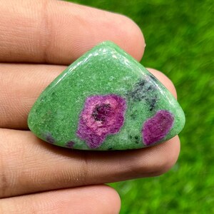 Puede incluir: Una piedra preciosa pulida de forma triangular que presenta una base de color verde vibrante con dos inclusiones de color magenta distintas e irregulares. La superficie de la piedra es lisa y brillante, mostrando una textura natural moteada en toda la matriz verde.
