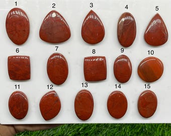 May include: A collection of fifteen polished, reddish-brown jasper cabochons arranged in three rows on a white display board. Each stone is numbered from 1 to 15 and features unique natural patterns, smooth surfaces, and various geometric shapes.