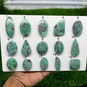 Può includere: Una collezione di quindici cabochon lucidi di rubino in zoisite disposti su un pannello bianco, numerati da 1 a 15. Ogni pietra presenta un motivo unico di matrice di zoisite verde brillante con inclusioni di rubino color magenta.