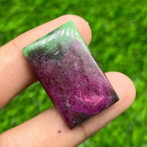 Op de afbeelding: Een rechthoekige cabochon edelsteen met een levendig magenta en dieppaars midden dat overgaat in een helder limoengroen gedeelte aan de bovenrand. De steen heeft een gepolijst, glad oppervlak met zichtbare natuurlijke minerale insluitingen.