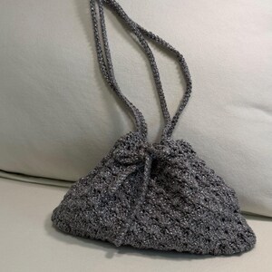 May include: A small, metallic silver drawstring pouch crafted from crocheted yarn. The bag features a textured, diamond-like pattern throughout its body and includes two long, thin crocheted straps that serve as both a closure and a handle.