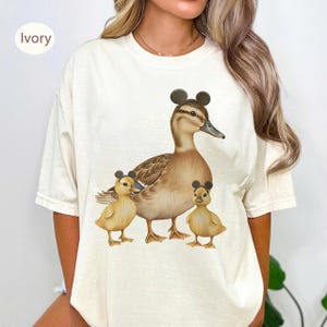 Pode incluir: Uma t-shirt cor de marfim com uma ilustração gráfica de um pato grande castanho e dois patinhos amarelos. Cada um dos três patos usa um par de orelhas de rato circulares pretas na cabeça. A t-shirt tem um decote redondo.