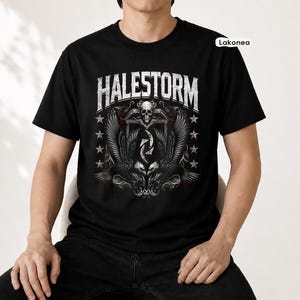 Op de afbeelding: Een zwart T-shirt met korte mouwen en een grafische print met de witte tekst HALESTORM. Het ontwerp bevat een centrale schedel, twee symmetrische adelaars met gespreide vleugels en een reeks witte sterren aan beide zijden van de afbeelding.