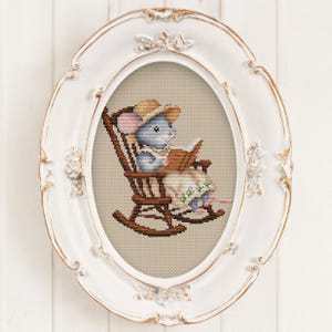 May include: A cross-stitch embroidery featuring a blue mouse wearing a straw hat and a floral dress, sitting in a brown wooden rocking chair while reading a book. The artwork is displayed within an ornate, oval-shaped white and gold frame.
