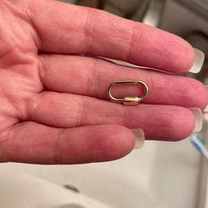 Könnte beinhalten: Ein kleiner, ovaler Federring-Verschluss aus goldfarbenem Metall. Das Schmuckelement verfügt über eine glatte, polierte Oberfläche mit einem funktionalen Federmechanismus an einer Seite, der zum Verbinden von Ketten oder Anhängern bei der Schmuckherstellung dient.