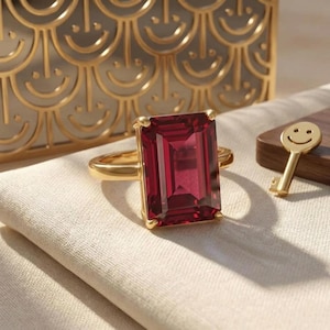 May include: A gold ring featuring a large, rectangular, deep red gemstone set in a four-prong setting. The ring is positioned on a light-colored fabric surface, with a small gold key charm and a decorative gold screen visible in the background.