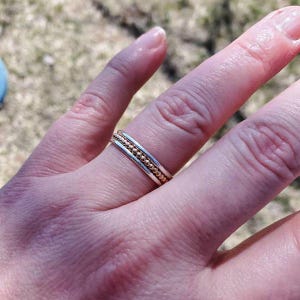 以下が含まれることがあります： 中央に小さな金色の金属ビーズのバンドがあり、その両側を滑らかな銀色のバンドが挟んでいる指輪。3つの明確な平行な列で構成されており、指の上で質感のある層状の外観を作り出している。