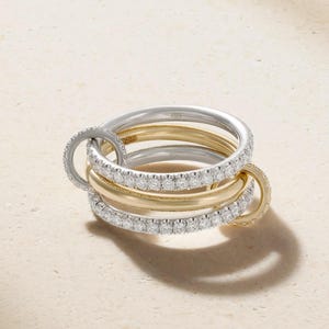 May include: A multi-layered ring set featuring two smooth gold bands and two silver-toned bands encrusted with small clear stones. Two smaller circular rings are linked through the main bands, creating a complex, interconnected design.
