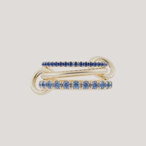 May include: A gold ring featuring three interconnected bands. The top band is set with small dark blue stones, the middle band is a plain polished gold hoop, and the bottom band is set with larger, lighter blue stones in a channel setting.
