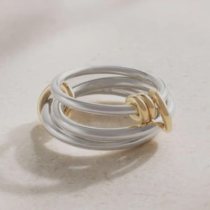 May include: A multi-band ring featuring three interlocking silver-toned bands and one smaller gold-toned band. The bands are smooth and circular, with the gold-toned band looped through the silver-toned bands to create a connected, layered design.