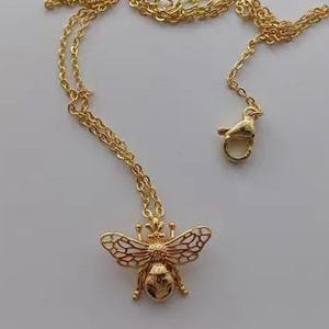 Könnte beinhalten: Eine goldfarbene Halskette mit einem Bienenanhänger, der weiße Emaille-Details auf den Flügeln aufweist. Der Anhänger ist an einer zarten goldfarbenen Kette befestigt, die einen Karabinerverschluss für einen sicheren Halt am Hals besitzt.