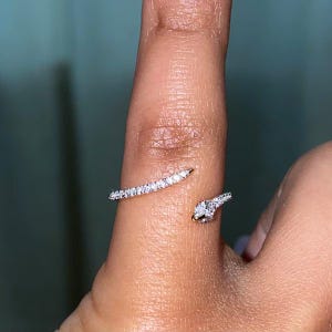 以下が含まれることがあります： 小さな透明な石が埋め込まれた曲線状のバンドと、反対側の端にある単一の大きな丸い透明な石を特徴とするオープンタイプのゴールドリングで、指を部分的に包み込むバイパスデザインになっています。