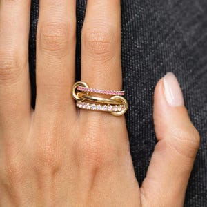 May include: A gold ring featuring three interconnected bands. The top band is set with small pink stones, the middle band is a plain polished gold loop, and the bottom band is set with small clear white stones, creating a layered appearance.