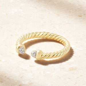 Può includere: Un anello aperto in oro caratterizzato da una texture a cavo intrecciato lungo la fascia. Le due estremità dell'anello sono rifinite con dettagli in oro liscio, ciascuno incastonato con un gruppo di piccole pietre rotonde, chiare e luminose.