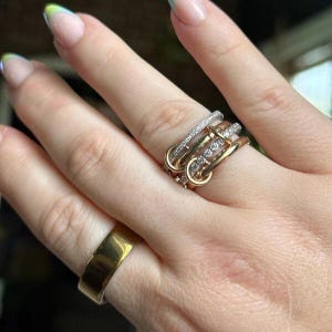 以下が含まれることがあります： ゴールドとシルバーのトーンが混ざり合った3つのインターロッキングリングのスタック。1つはシンプルなゴールドバンド、もう1つは小さなクリアストーンがセットされたゴールドバンド、3つ目はクリアストーンがパヴェセットされたシルバー調のバンドです。