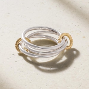 May include: A multi-band ring featuring three smooth silver bands intertwined with two smaller, textured gold bands. The silver bands are stamped with the letters SK and the number 925, indicating sterling silver composition. The gold bands feature small, clear stones.