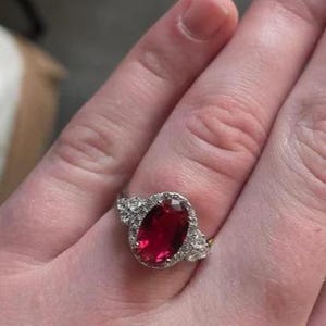 Könnte beinhalten: Ein silberfarbener Ring mit einem großen, oval geschliffenen, tiefroten Edelstein in der Mitte. Der zentrale Stein ist von einem Kranz aus kleinen, klaren, funkelnden Steinen umgeben, mit weiteren klaren Steinen entlang der Ringschiene.