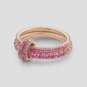 Puede incluir: Un anillo de oro rosa con un diseño de doble banda conectado por un eslabón circular. Toda la superficie del anillo está incrustada con pequeñas piedras preciosas redondas de color rosa vibrante que cubren las bandas y el eslabón central.