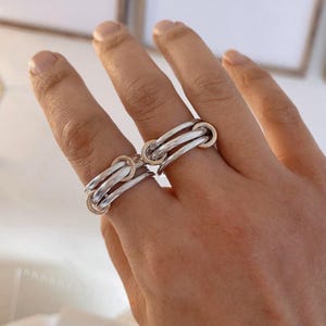 Op de afbeelding: Twee ringen gedragen aan aangrenzende vingers, elk opgebouwd uit meerdere in elkaar grijpende banden. De ringen combineren gepolijst zilverkleurig metaal met kleinere, roségoudkleurige ronde verbindingsstukken die de afzonderlijke banden verbinden.