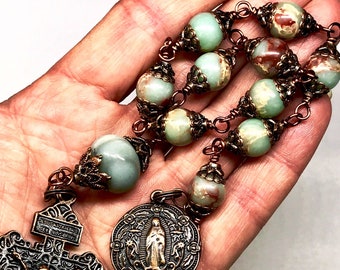 Puede incluir: Un rosario envuelto en alambre de cobre con cuentas de ágata verde y marrón, un crucifijo de cobre con una figura de Jesucristo y un medallón de cobre con una imagen de la Virgen María.