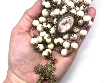 May include: White rosary beads with a brass crucifix and a brass oval pendant featuring a religious image. The rosary is held in a hand.